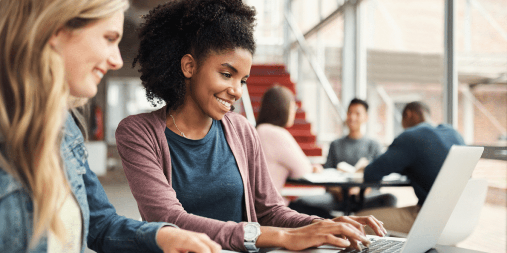 Two female college students sitting in common area on campus looking at laptop.