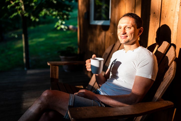 Man happy sitting relaxing on rocking chair lounge on porch of house in morning wooden cabin cottage drinking coffee or tea from cup mug