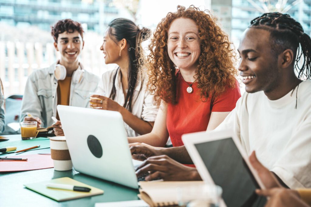 Multiracial university students sitting together at table with books and laptop - Happy young people doing group study in high school library - Life style concept with guys and girls in college campus