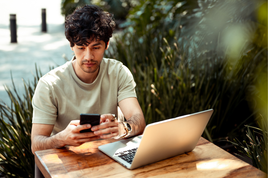 Young man sitting outside at a table with his laptop and looking at his phone.