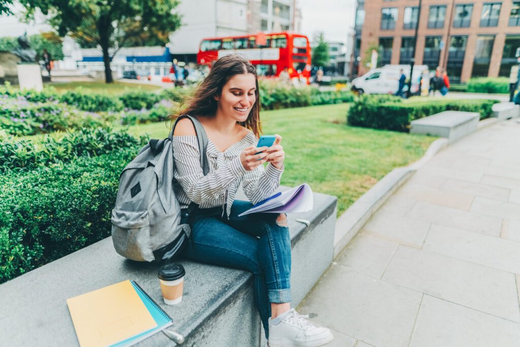 Young girl sitting on bench and using smartphone to see how soon she can refinance her student loans.