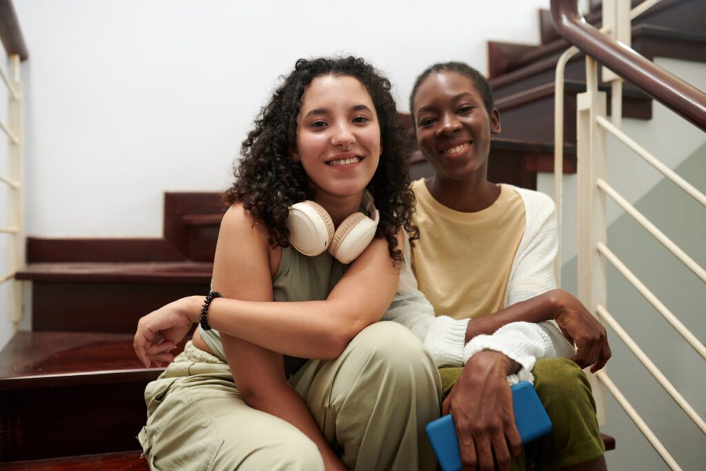 Joyful teenage friends sitting on steps in rented house and smiling at camera