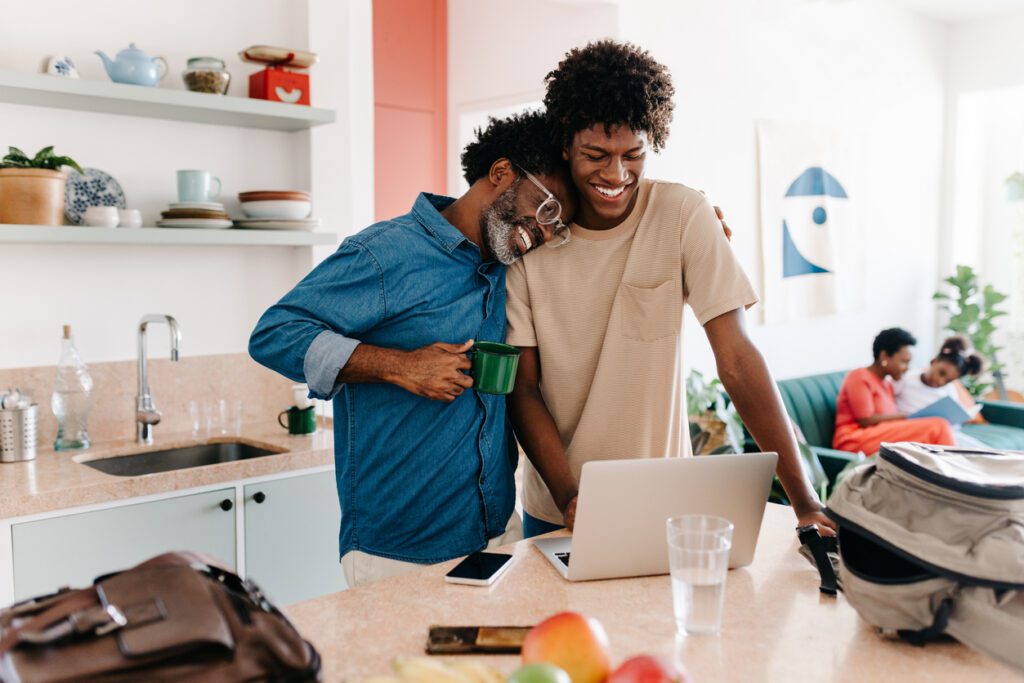 Mature black dad and his teenage son share a loving moment in their kitchen as they use a laptop and prepare for their day ahead. Father and son enjoying family life at home.